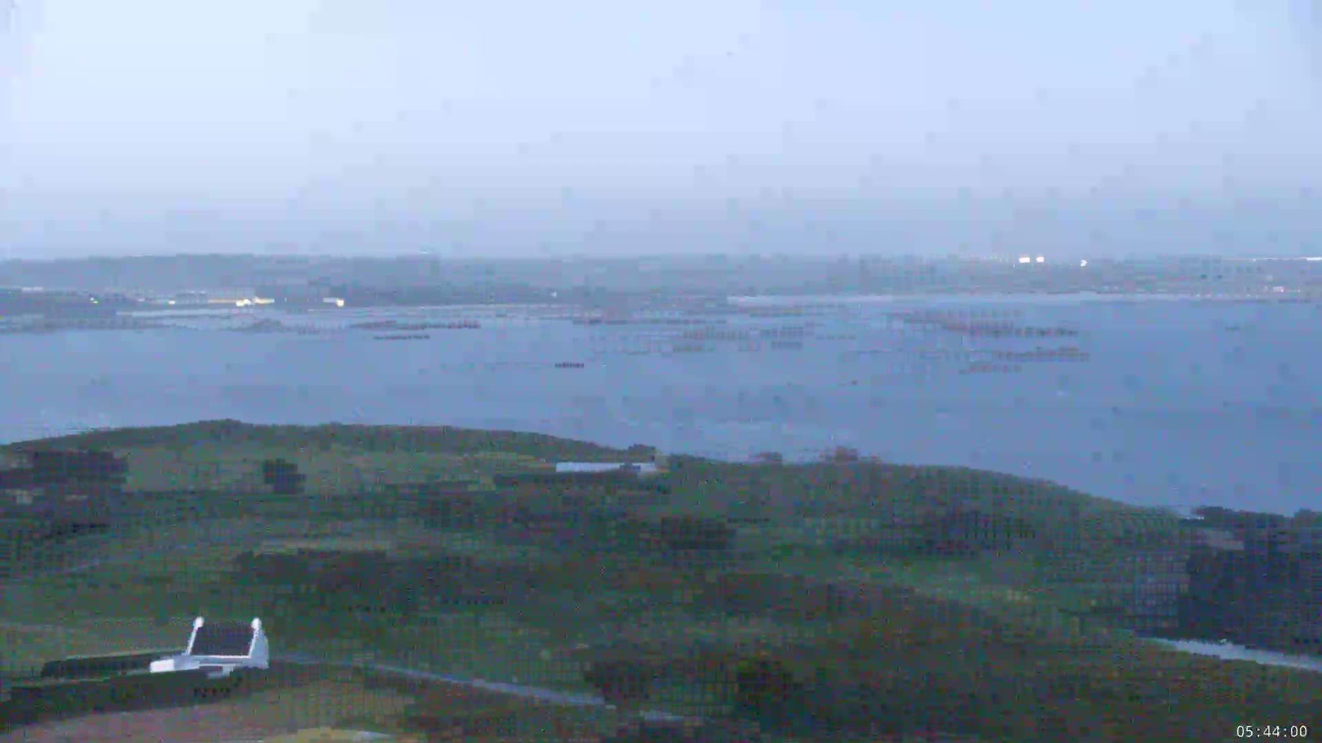 A hazy, overcast view shows a large body of water with a distant shoreline and a foreground of low-lying, green hills.