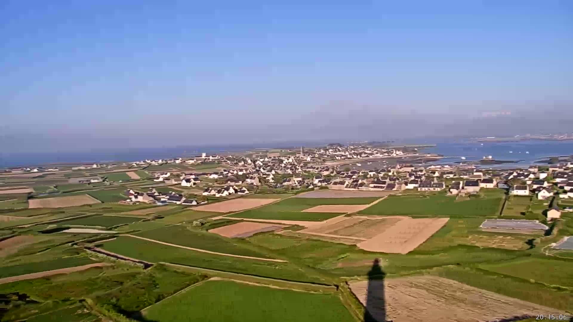 An elevated view shows a coastal town surrounded by agricultural fields under a clear, sunny sky, with a body of water visible in the background.