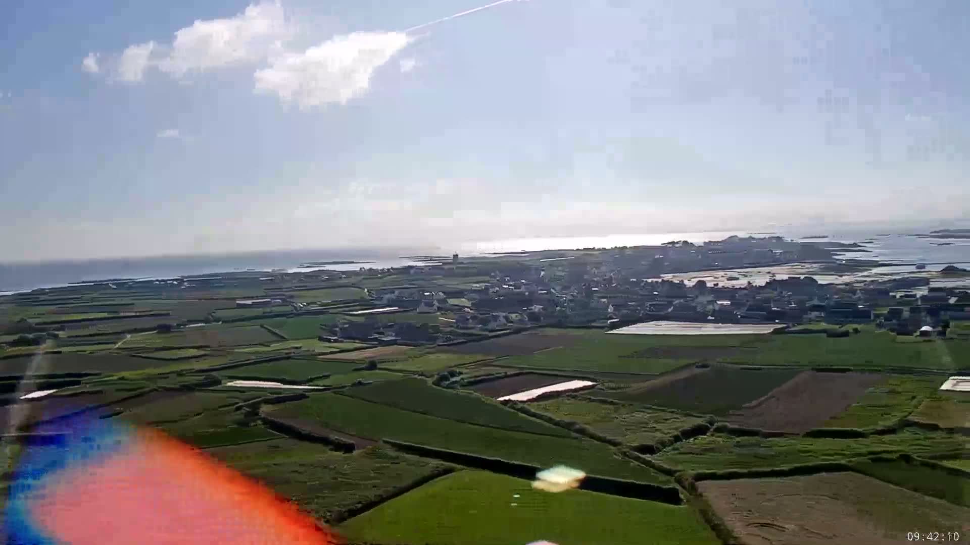 An aerial view shows a coastal town nestled amongst farmland under a partly cloudy sky.