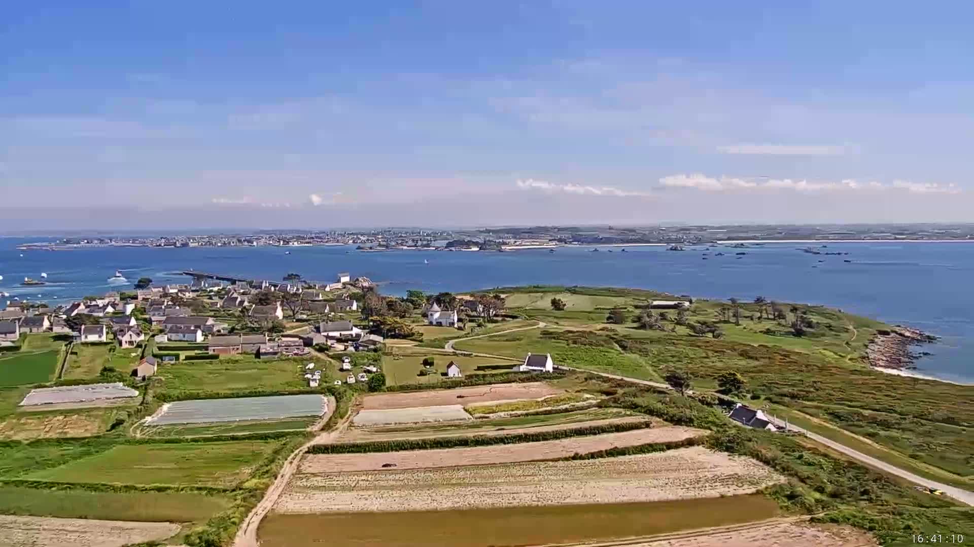 A small coastal village with farmland and a bridge connecting it to a larger town across the water, under a mostly clear blue sky.