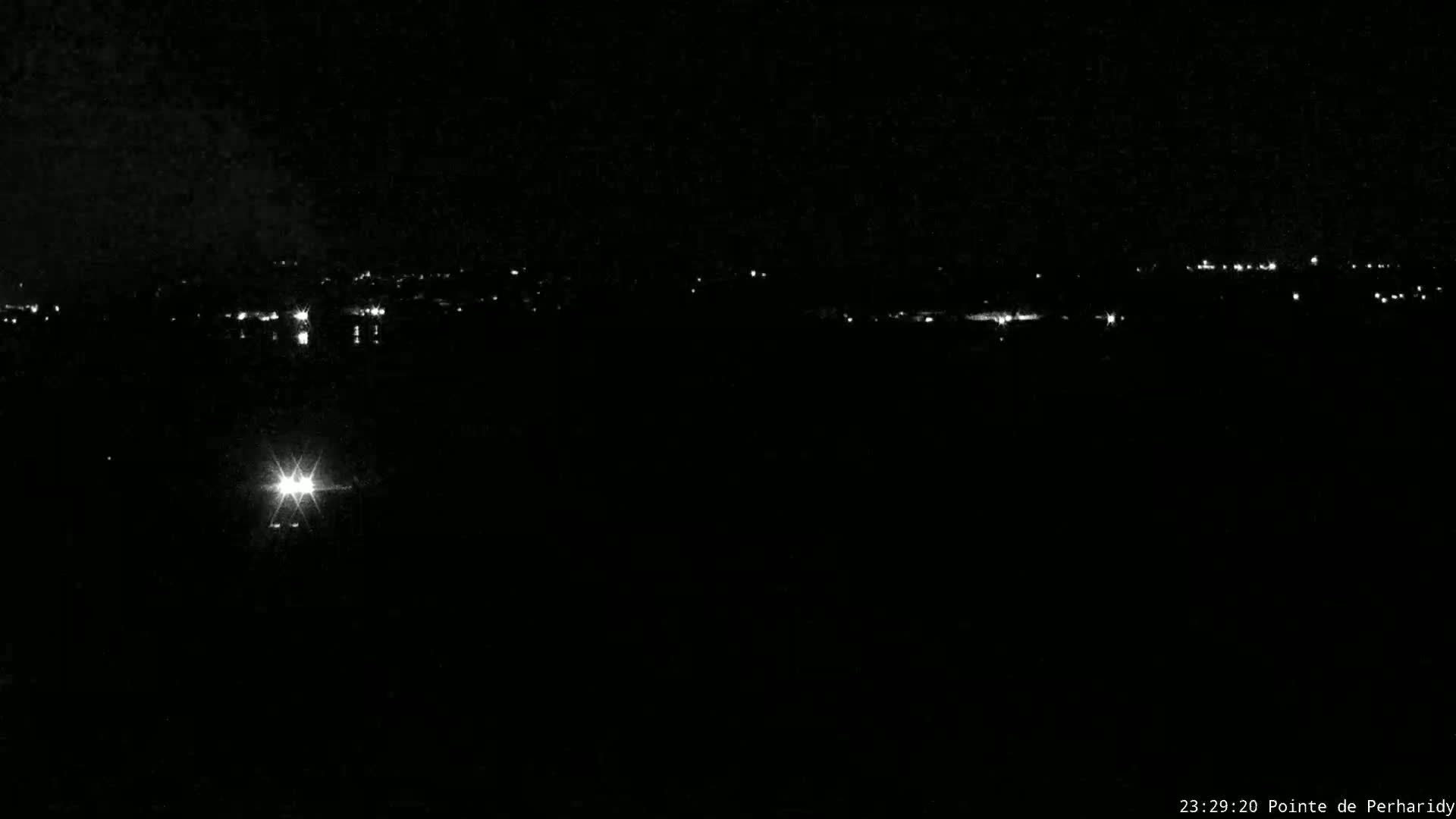 A nighttime long shot of a distant town's lights, with a single bright light source closer to the camera in the dark.