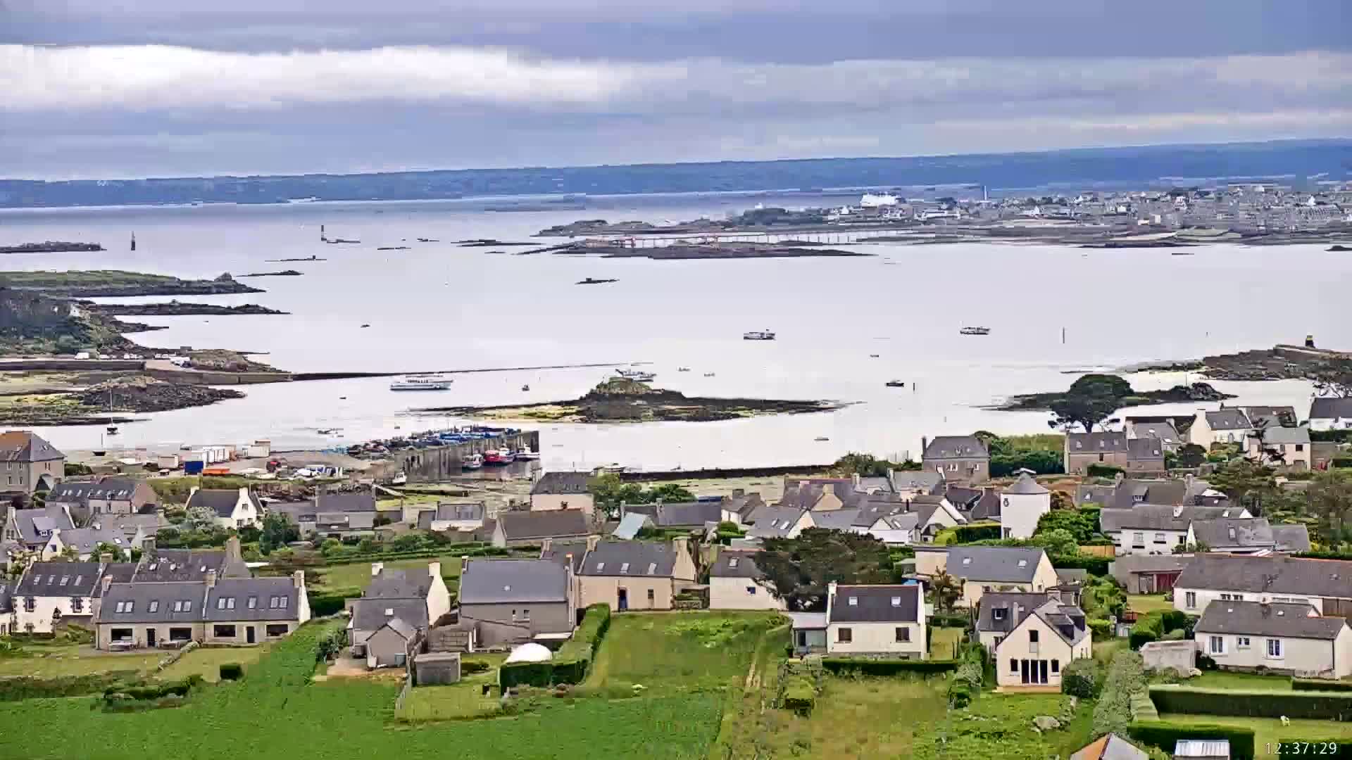 A coastal town with numerous houses sits beside a calm body of water under a cloudy sky, with a larger body of water and more distant land visible in the background.