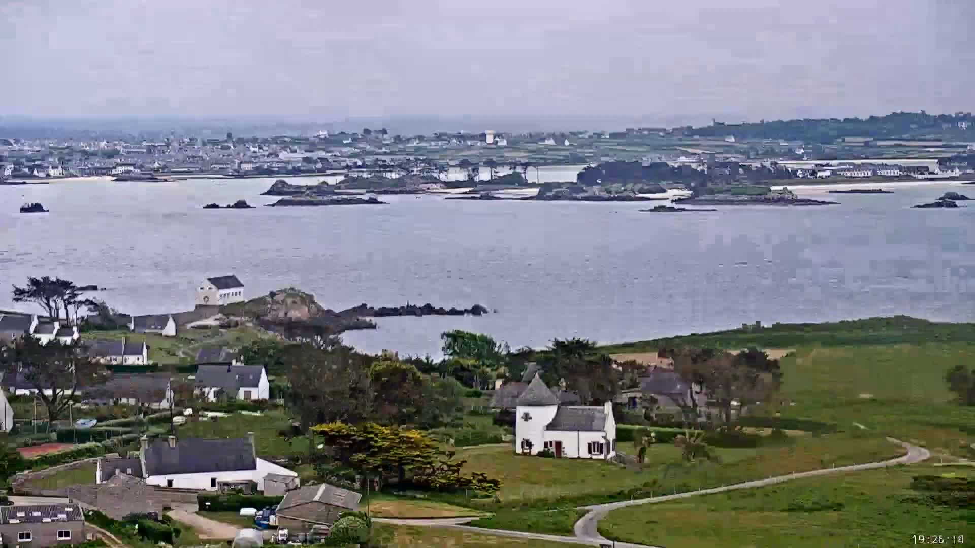 A coastal village with white houses and green fields overlooks a calm body of water under an overcast sky, with a larger town visible in the distance.