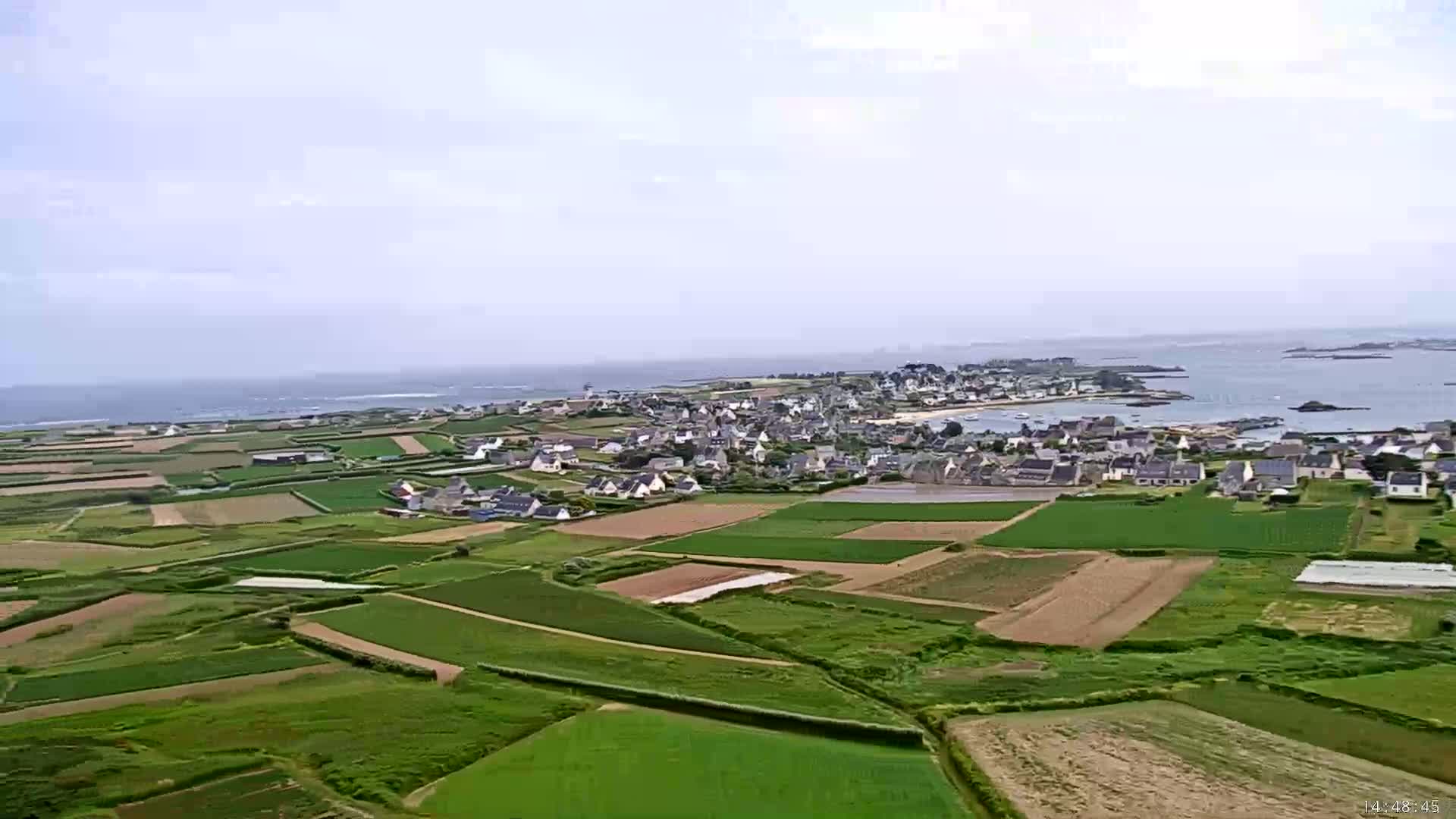 An aerial view shows a coastal village nestled amongst farmland bordered by a calm ocean under a mostly cloudy sky.