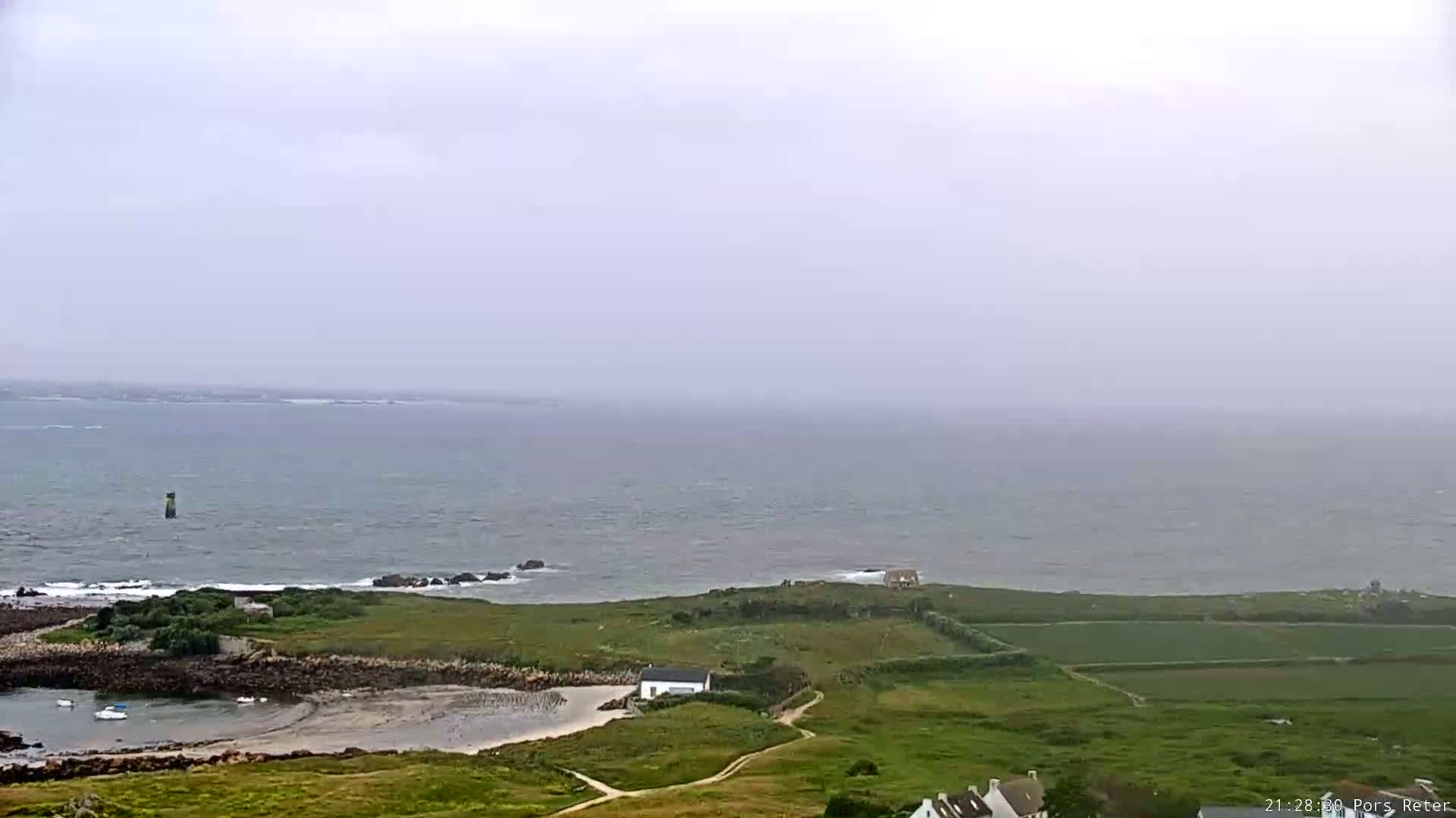 A hazy, overcast day reveals a coastal scene with a small, grassy island, a few buildings, and a calm sea extending to a distant horizon.