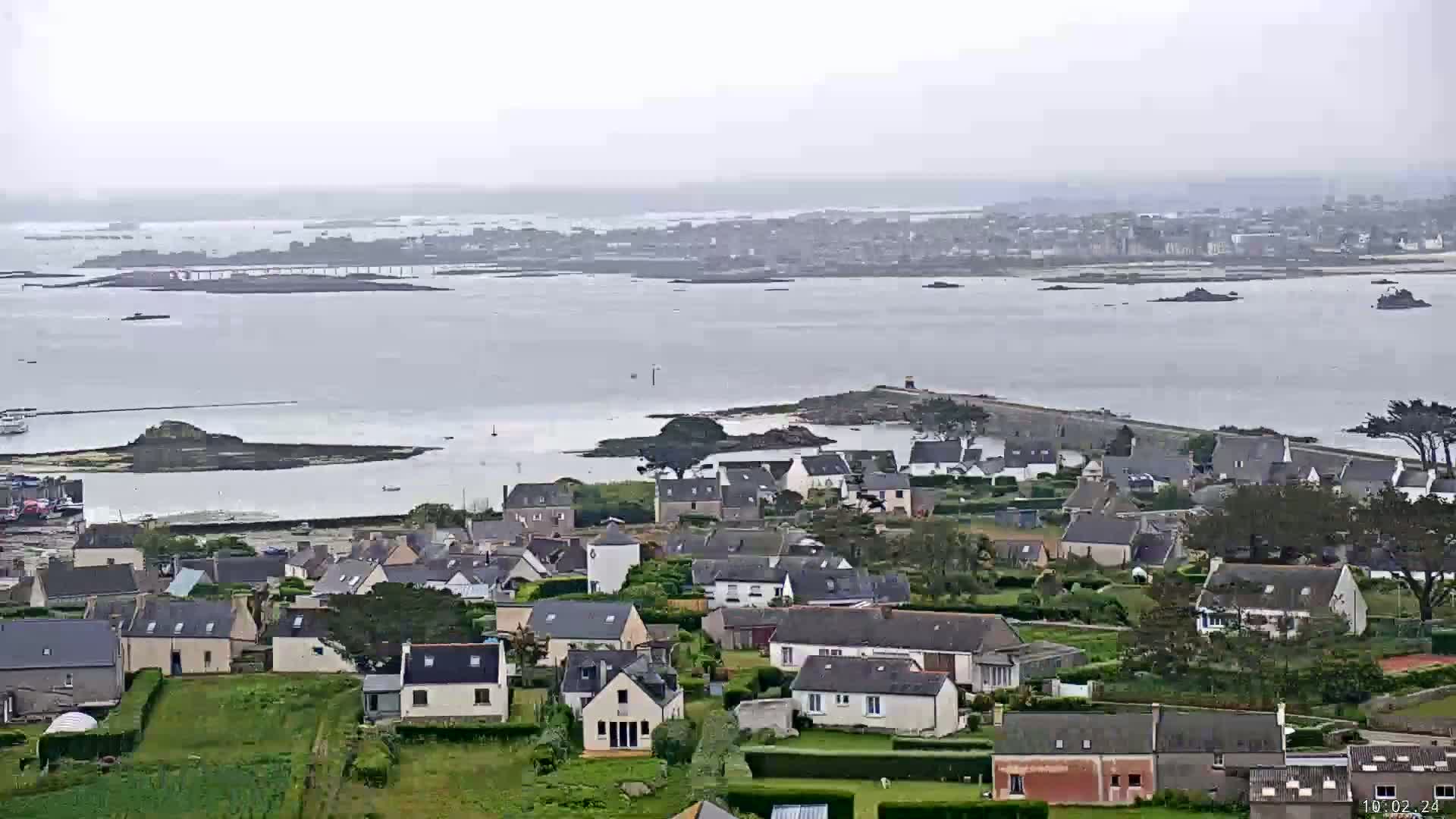 A coastal town with grey houses and green lawns overlooks a calm, grey sea under an overcast sky.