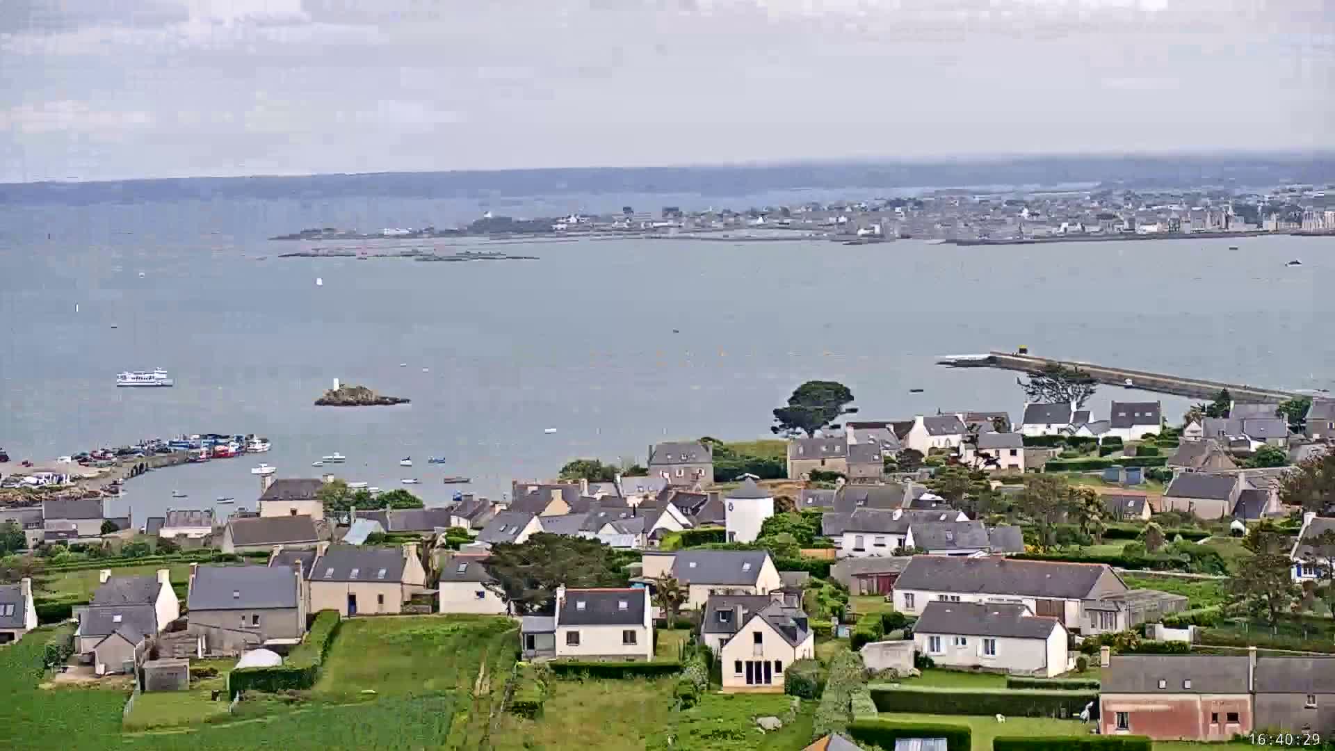 A coastal town with houses clustered together overlooks a calm bay, with a larger town visible in the distance under a partly cloudy sky.