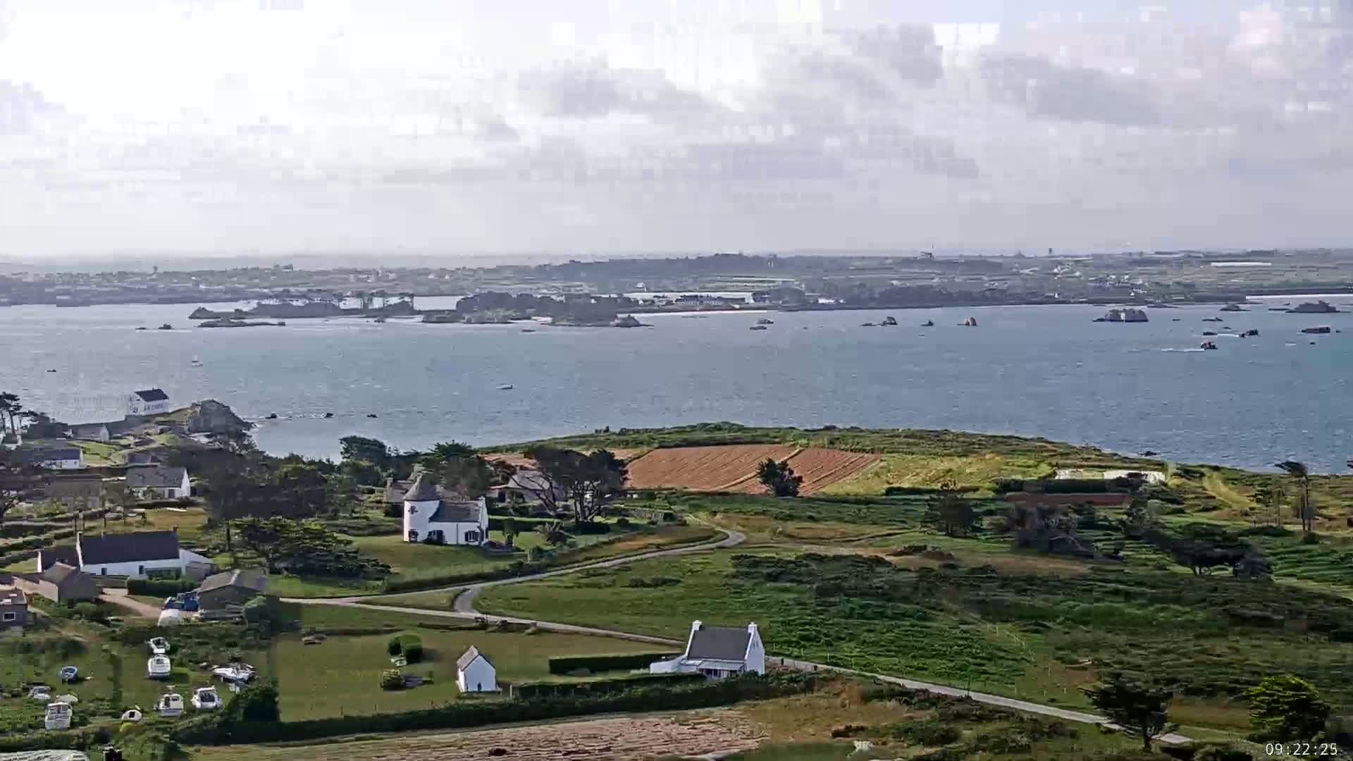 A coastal village with white houses and farmland overlooks a calm body of water dotted with small islands under a partly cloudy sky.