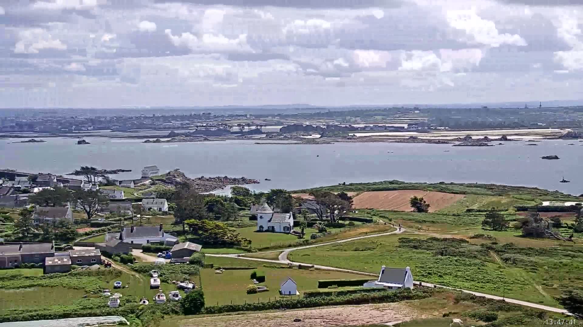 A coastal village with white houses and green fields is visible under a partly cloudy sky, overlooking a calm body of water with islands in the distance.