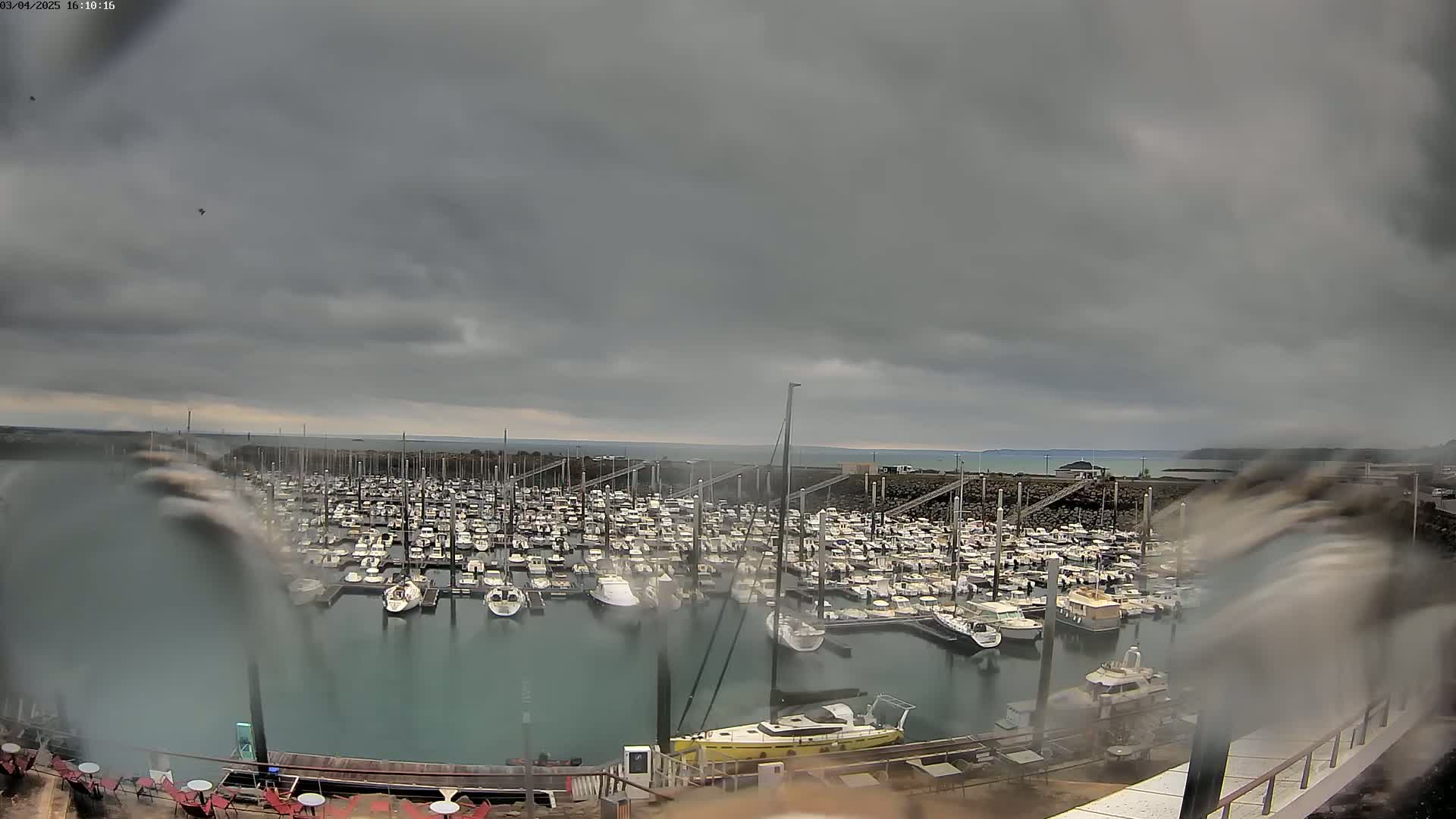 A large marina filled with many boats is seen under a cloudy, overcast sky, with rain or spray obscuring the view.