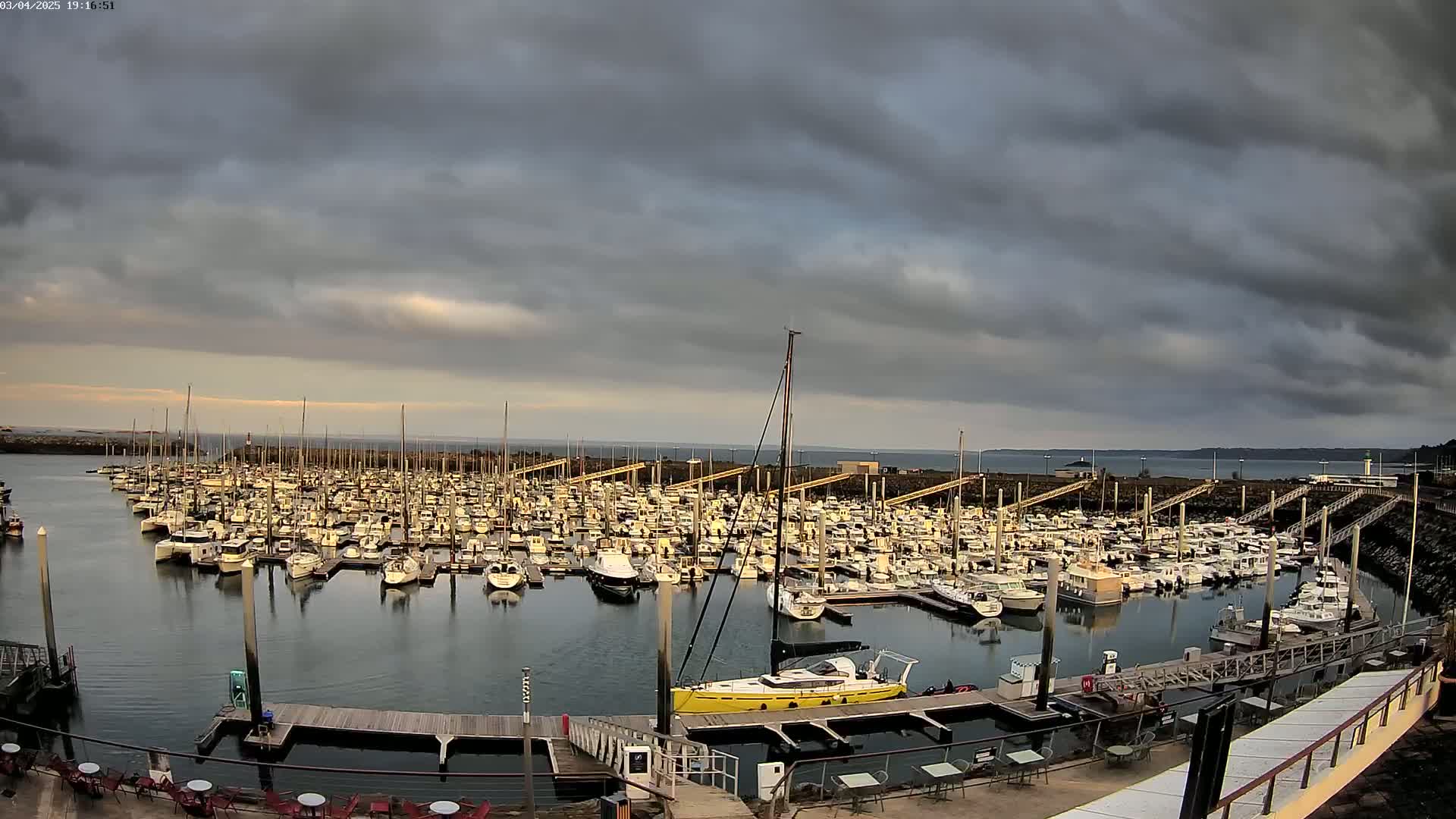 Many boats are docked in a marina under a cloudy sky.