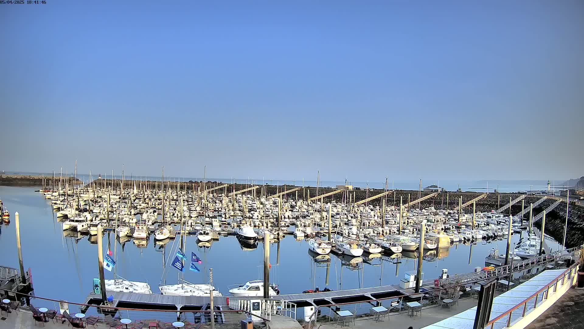 A calm, sunny day reveals a marina filled with numerous boats neatly docked, their reflections visible in the still water.