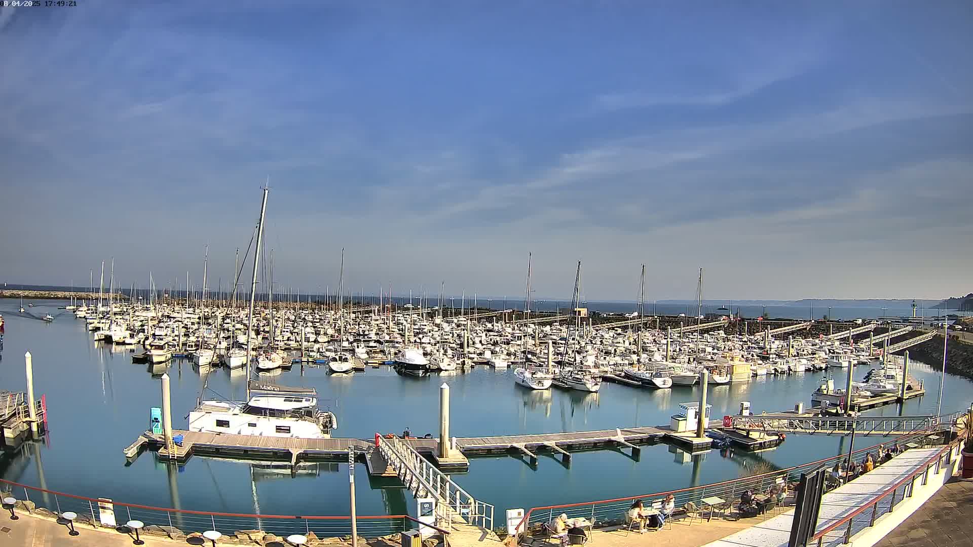 A large marina filled with numerous sailboats and motorboats is seen on a calm, sunny day with a clear blue sky.