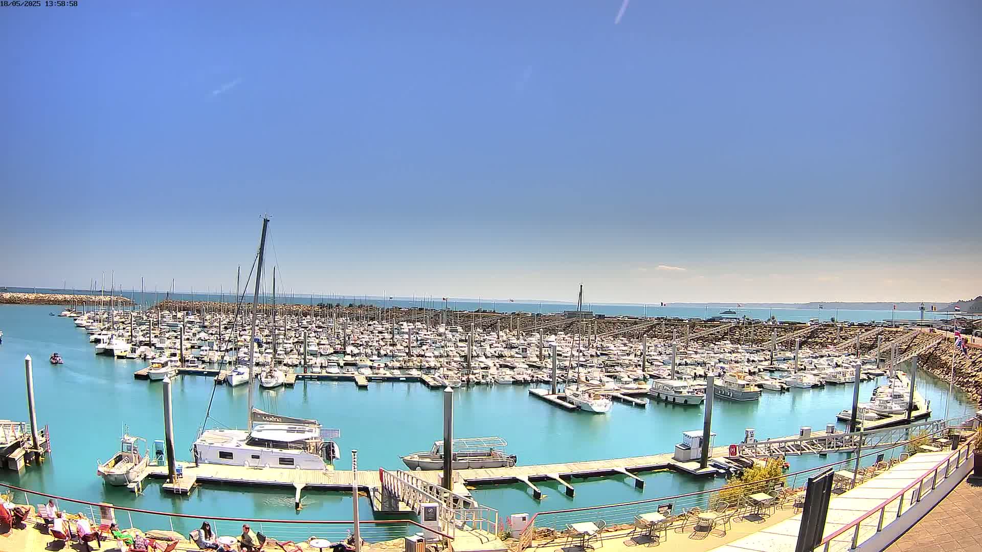 A large marina filled with numerous boats is seen on a sunny day with clear blue skies and calm turquoise water.