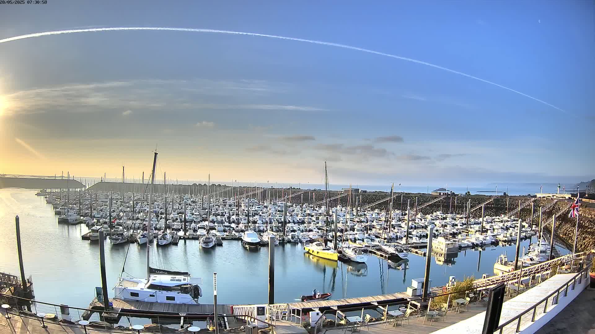 A calm, sunny day shows a large marina filled with numerous boats of various sizes, with a clear blue sky and a few thin clouds.