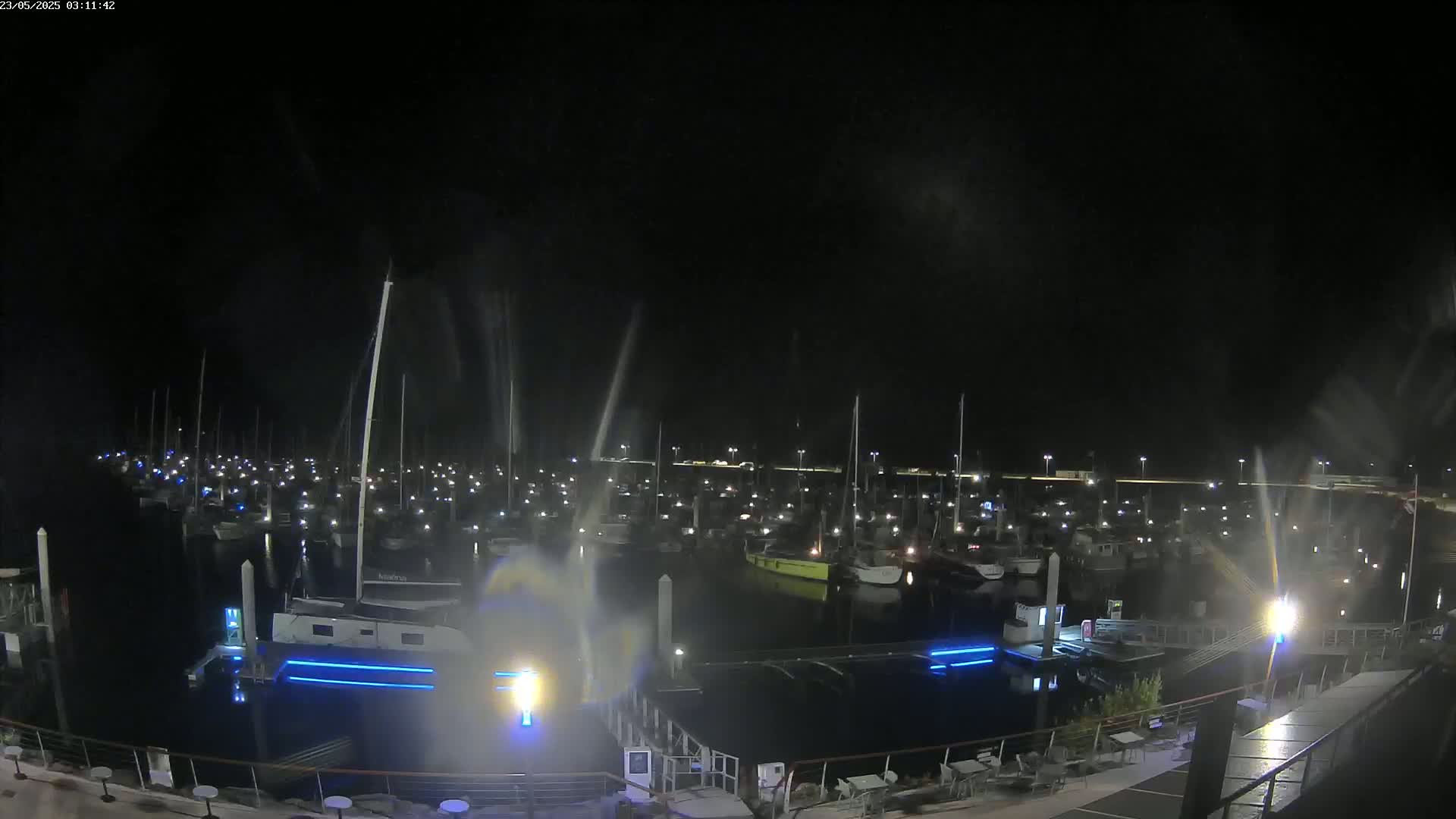 A nighttime view of a marina filled with boats, illuminated by lights, with some spray or mist visible in the air.