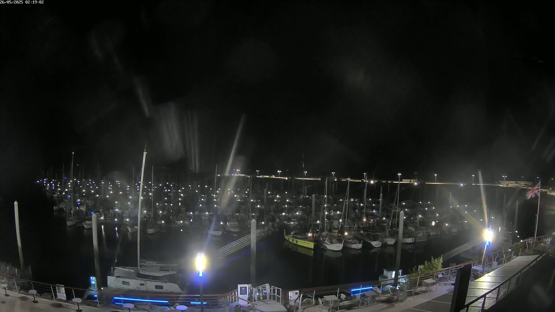 A nighttime view of a marina filled with numerous boats, illuminated by lights, under a dark, slightly hazy sky.