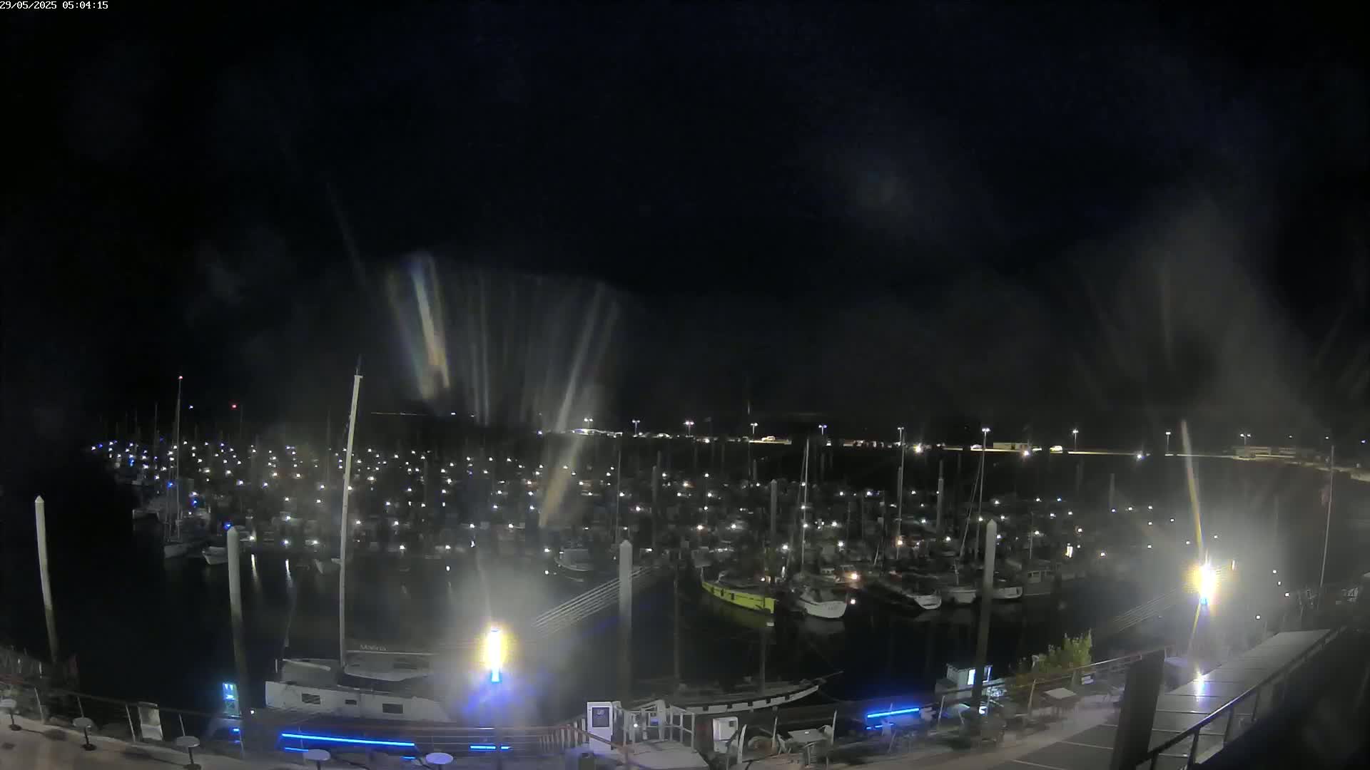 A nighttime view of a marina filled with numerous boats, illuminated by lights, under a dark, slightly hazy sky.