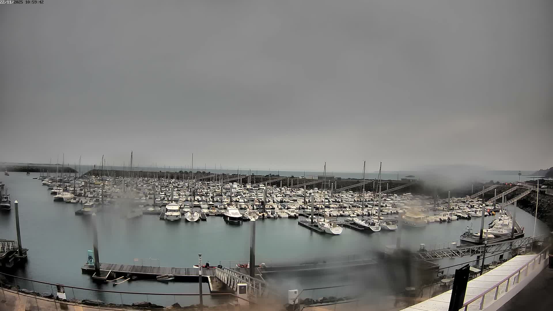 A large marina is densely packed with boats and sailboats under a dark, overcast sky with visible rain on the camera lens, suggesting wet and gloomy weather.