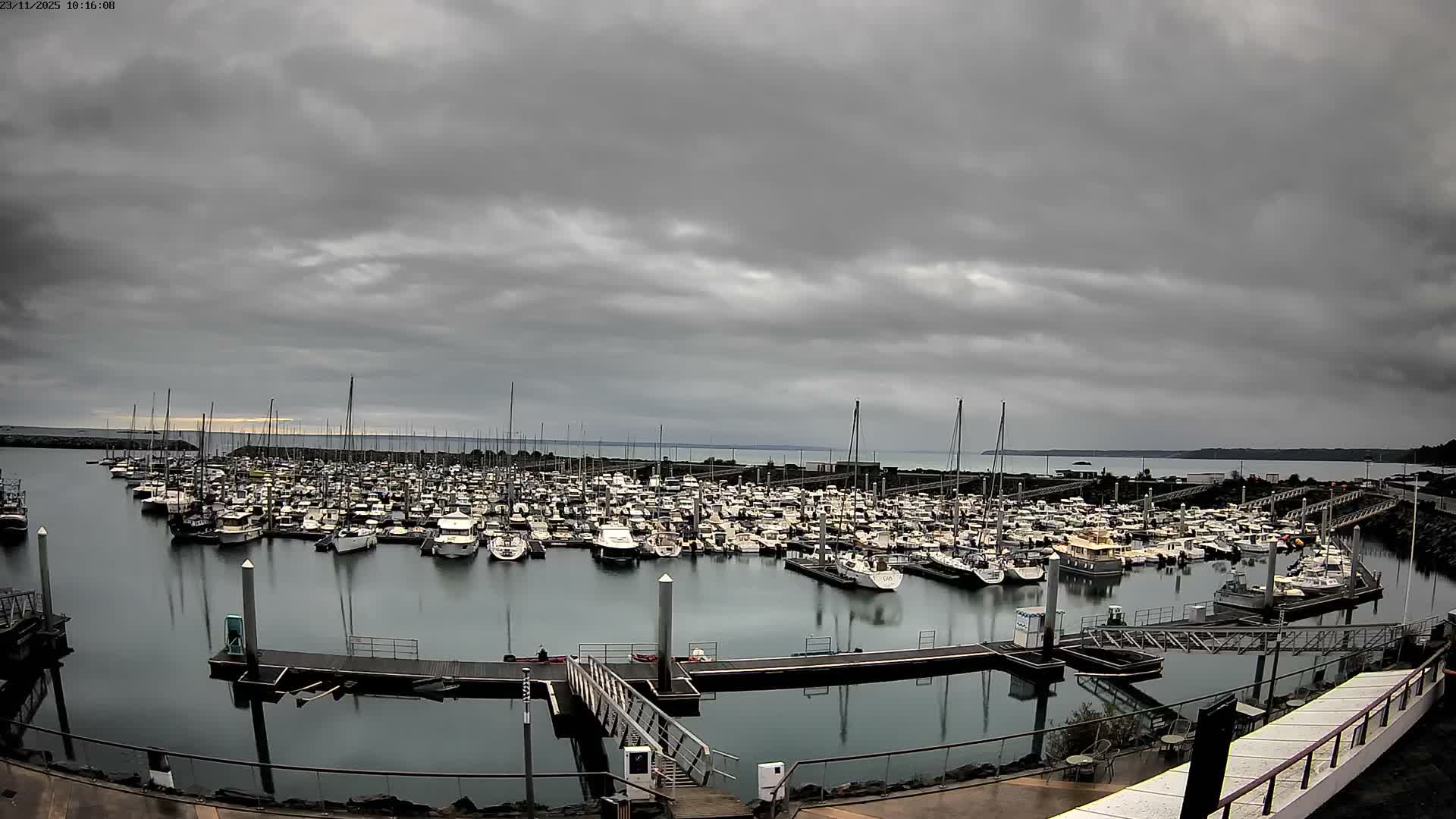 A marina is crowded with numerous boats and yachts under a heavily overcast and gloomy sky, with calm water reflecting the scene.