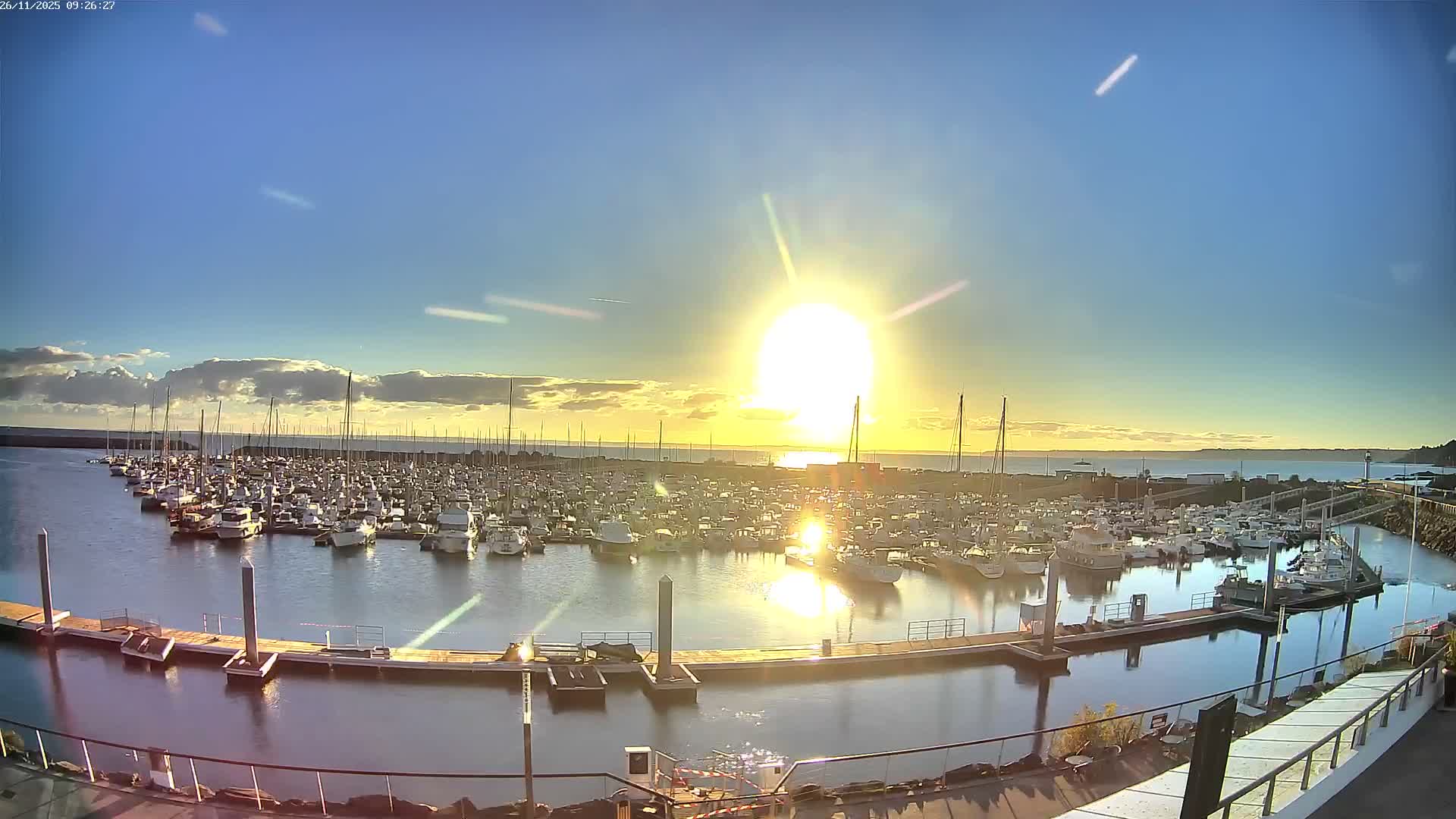 A sunny day illuminates a busy marina filled with numerous boats docked in calm water, with a bright low sun creating a strong glare and reflections across the scene under a clear to partly cloudy sky.