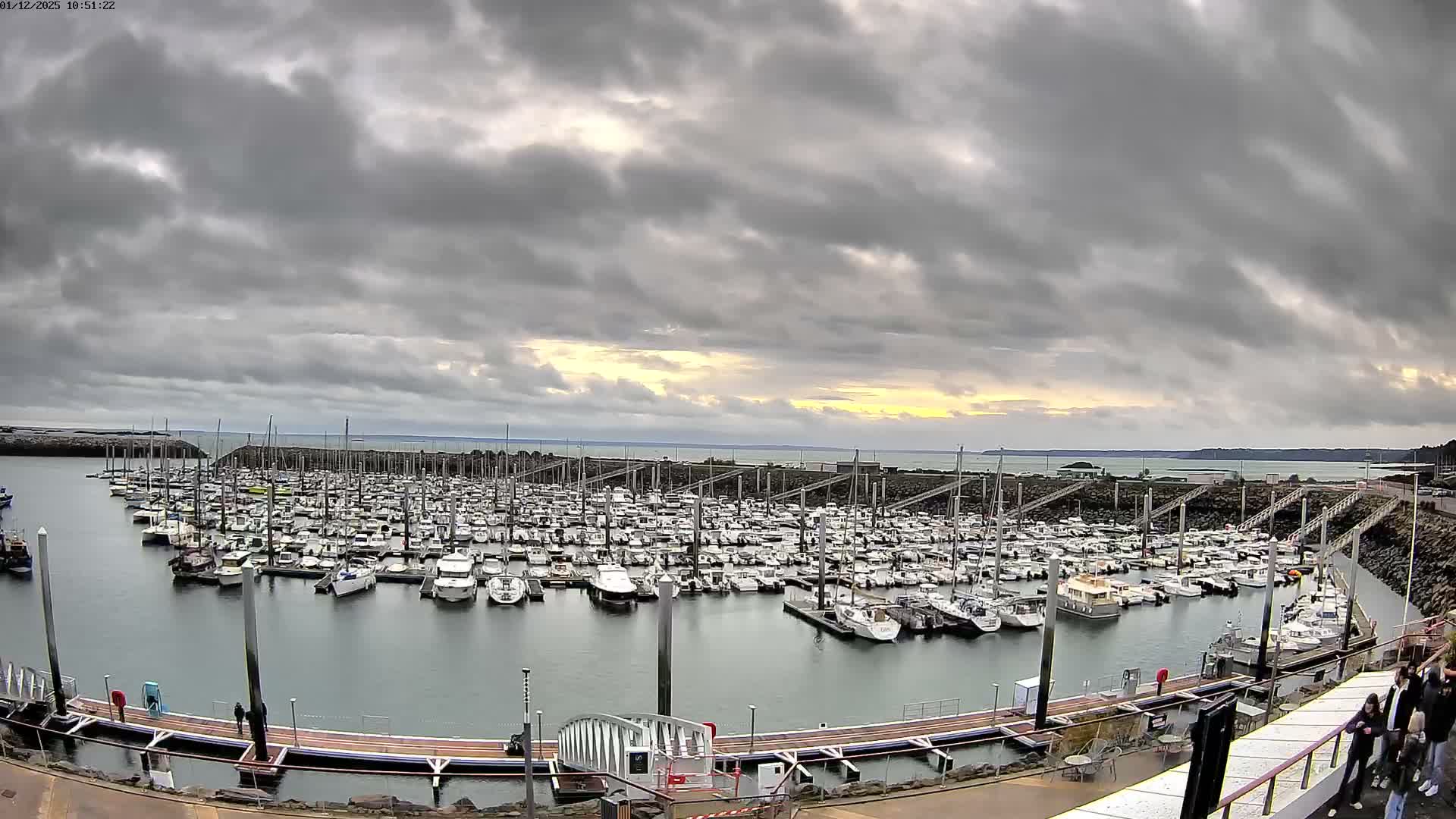 A vast marina filled with numerous docked boats is seen under a heavily overcast sky, with a sliver of golden light breaking through the clouds on the distant horizon.