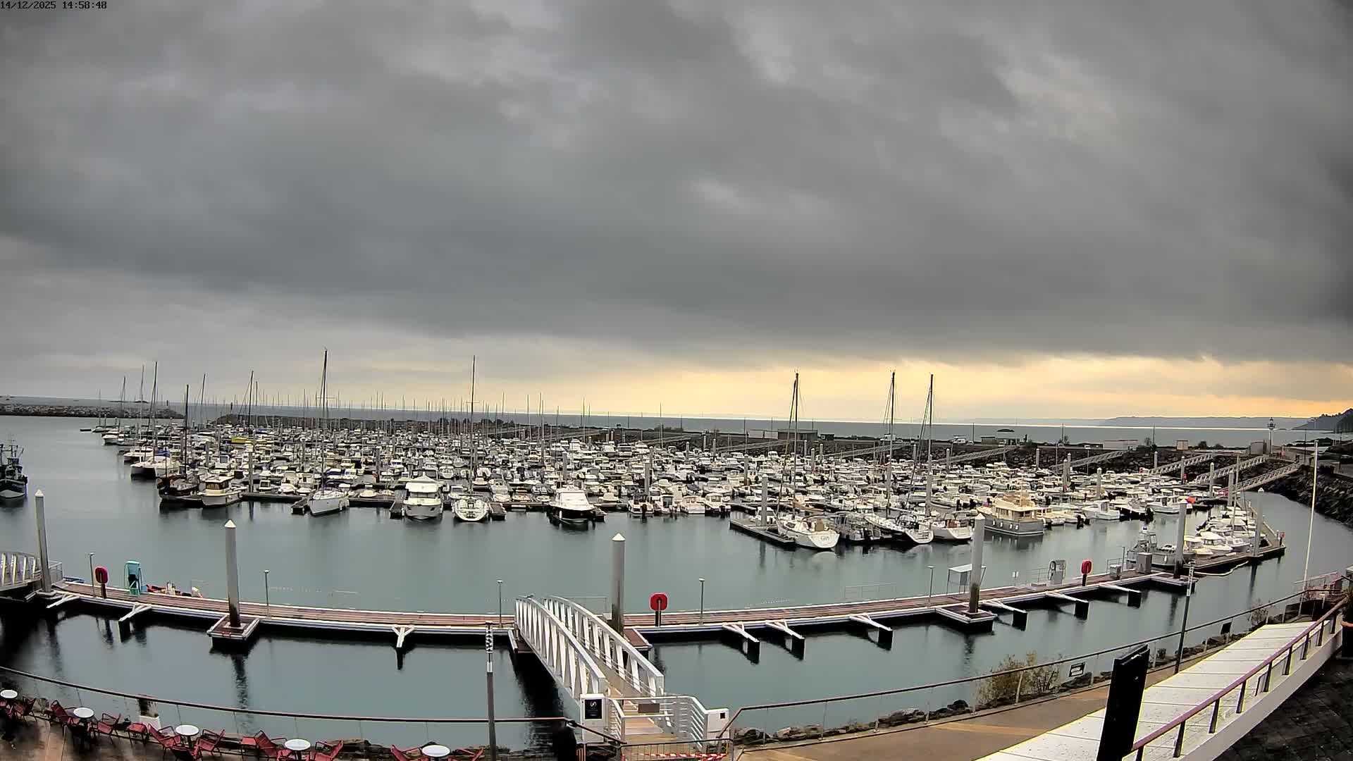 Numerous boats are docked in a calm marina under a clear, bright sky, with the sun low on the horizon casting a strong golden light across the water.
