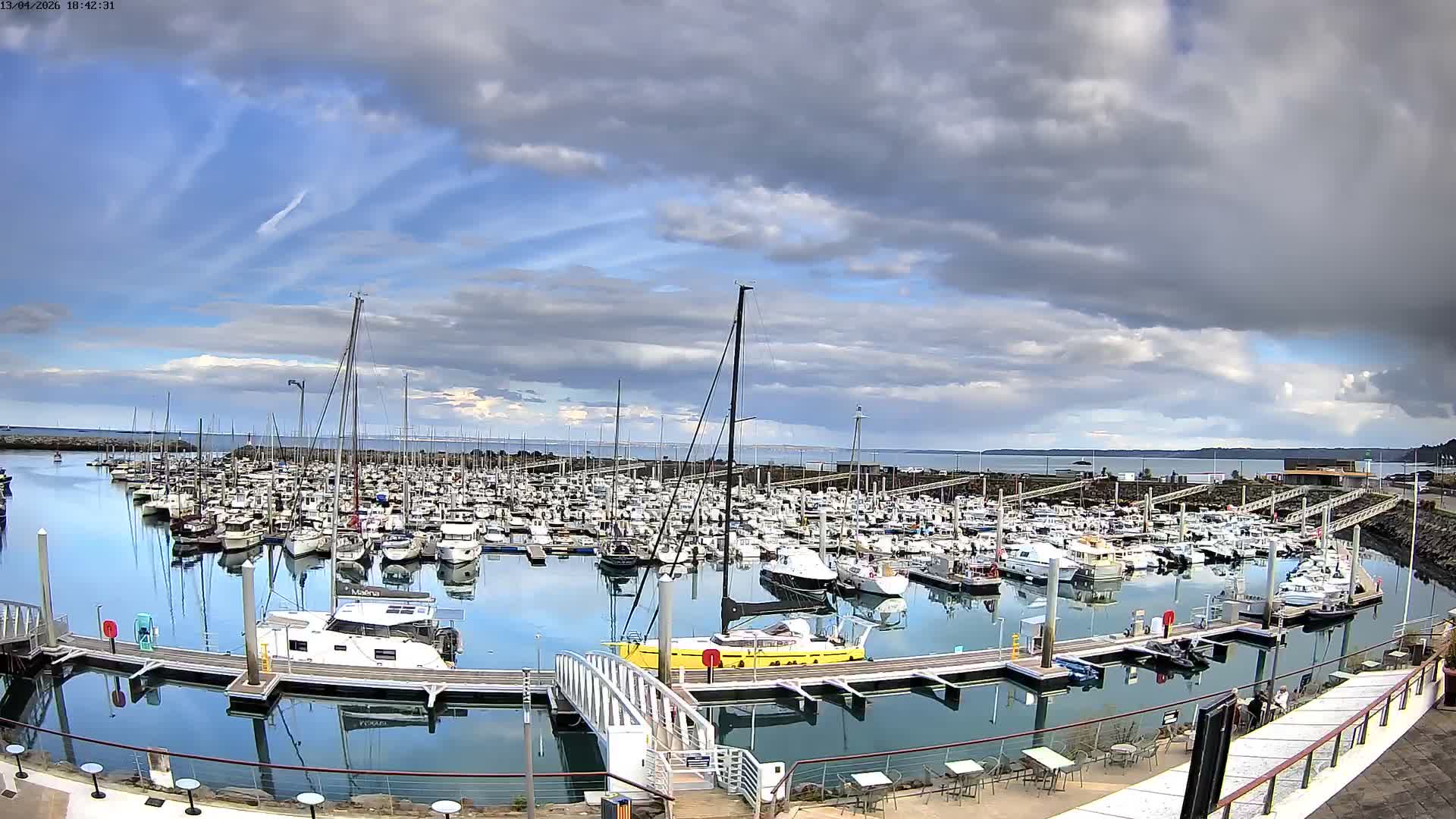 Numerous boats are docked in a calm marina under a clear, bright sky, with the sun low on the horizon casting a strong golden light across the water.