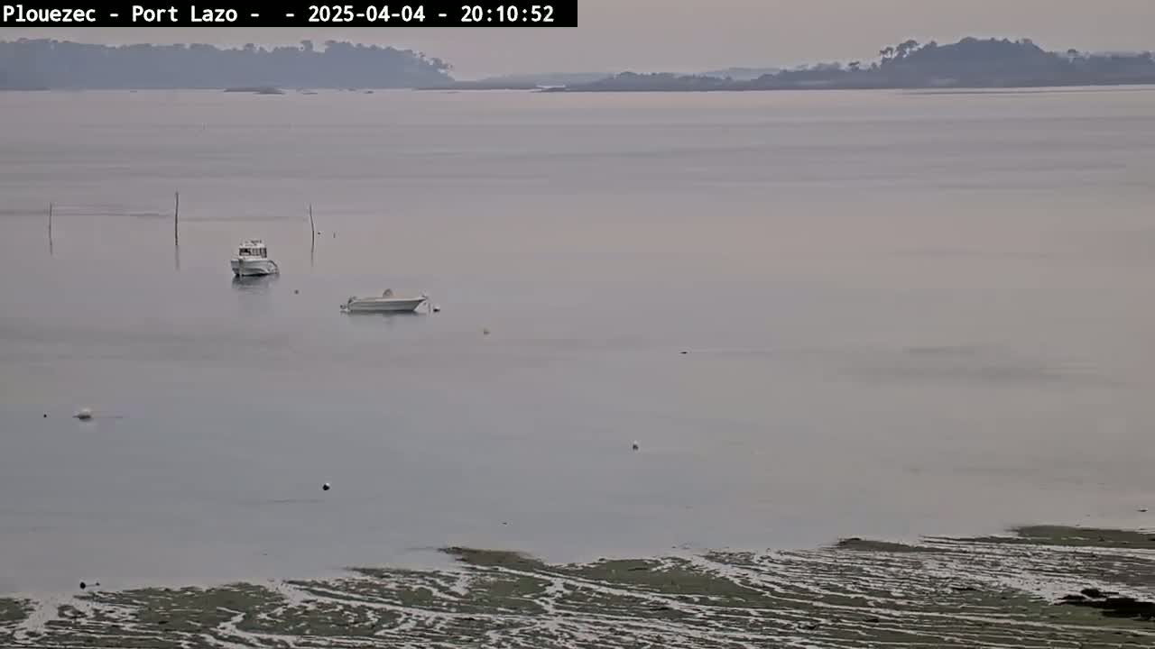 Two small boats are moored in calm, hazy water near a shoreline of exposed mudflats and a distant landmass.