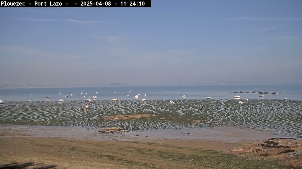 A mostly sunny day reveals a wide expanse of tidal flats patterned with rivulets, numerous small boats resting on the exposed seabed, and a calm body of water in the distance.