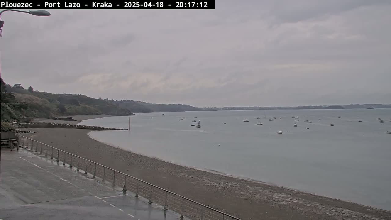 A mostly cloudy day shows a sandy beach, calm bay with several boats, and a grassy hill in the background.