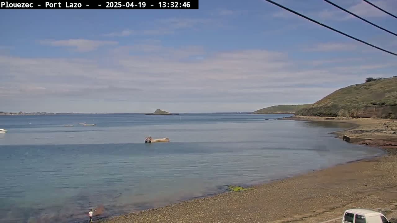 A calm, partly cloudy day shows a rocky beach with a few people and small boats on a bay, with islands in the distance.