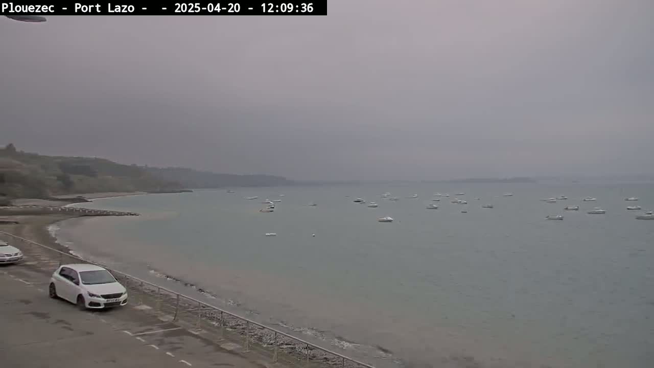 A gray, overcast day shows a sandy beach with several boats in the calm water, and two cars parked near the shoreline.