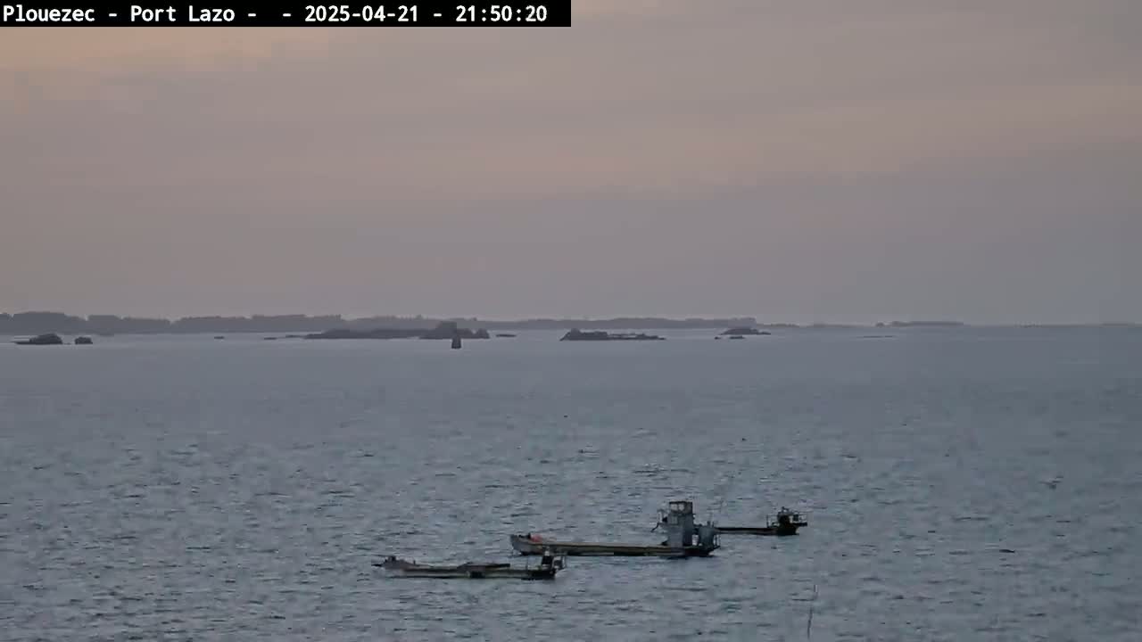 Three small boats are moored in calm, gray water under a dusky sky, with a distant, low-lying landmass visible on the horizon.