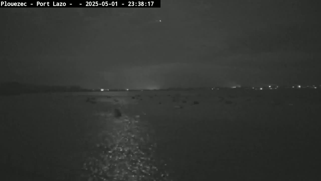 A nighttime, low-light image shows a dark body of water with faint reflections on its surface, and a distant shoreline with a few faint lights visible.