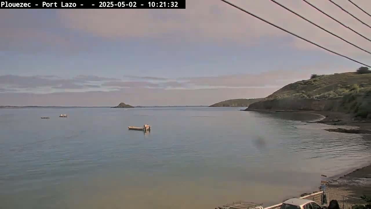 A calm bay with several small boats is visible under a mostly cloudy sky, with a small hill visible on the right.