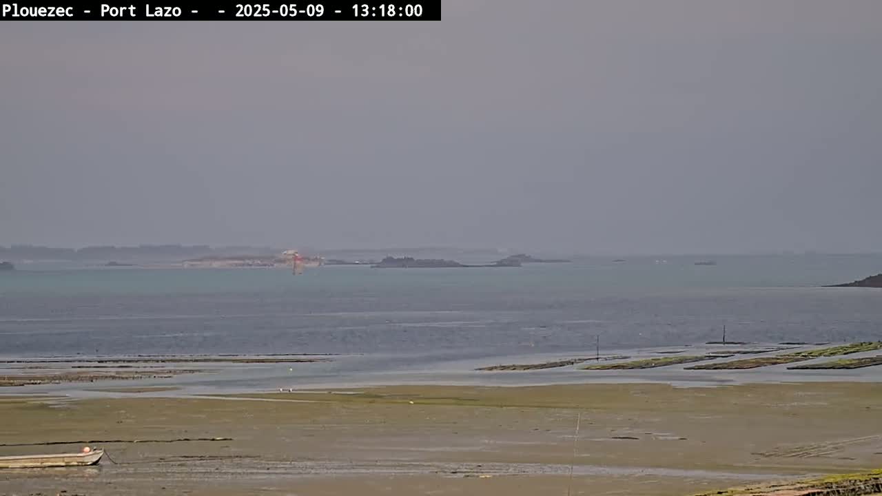 A hazy, calm day reveals a coastal scene with a small boat on a sandy shore near oyster beds, and a distant island visible across a calm body of water.