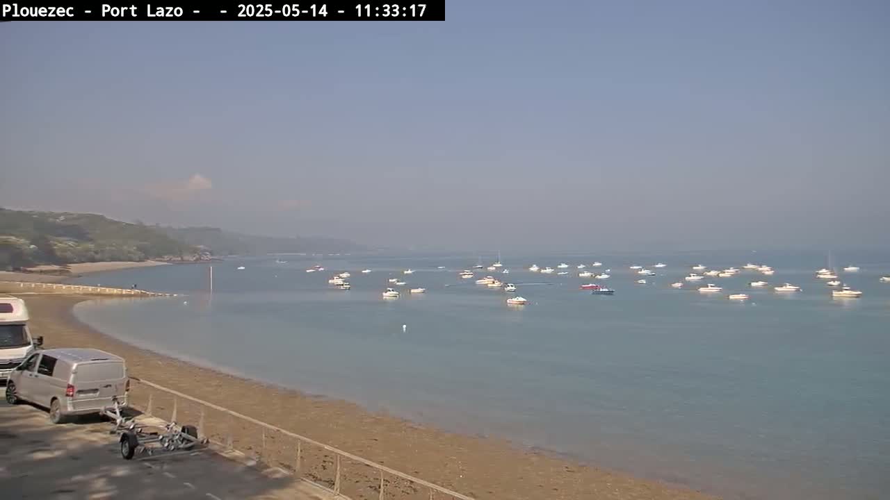 A hazy, sunny day shows a sandy beach with a campervan and boat trailer parked, overlooking calm water with numerous small boats at anchor.