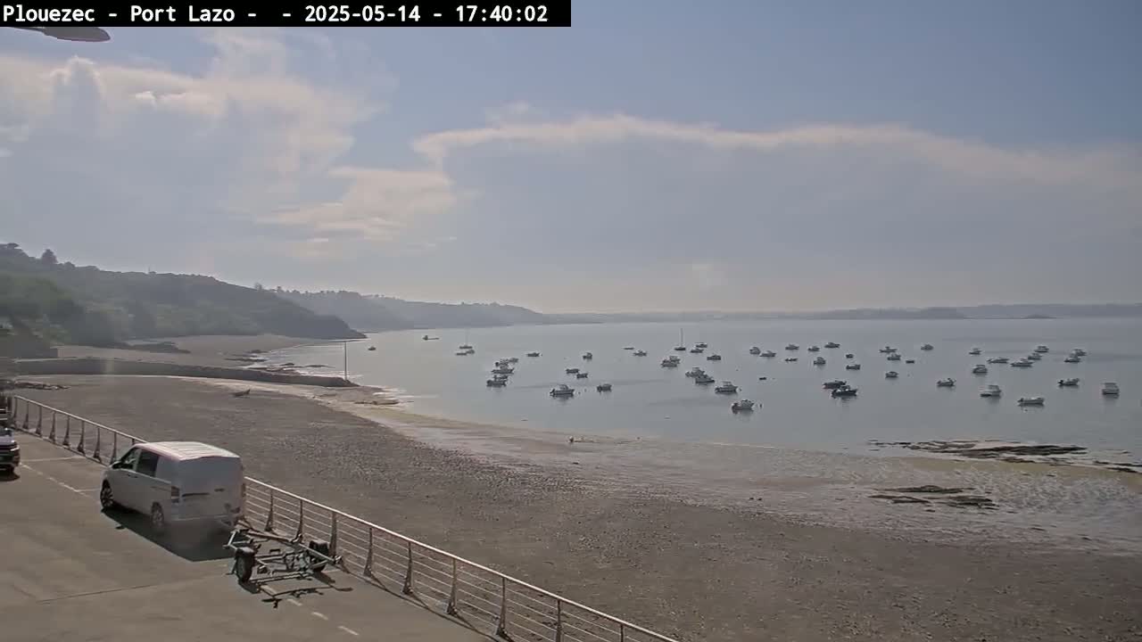 A sandy beach with a few boats in the calm water and a van parked nearby, under a mostly clear, bright sky.