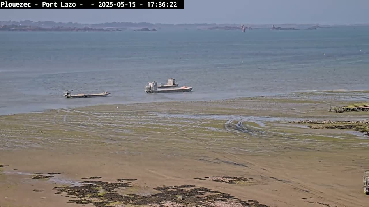 Two barges are moored in calm water near a sandy shore exposed at low tide, under a sunny sky.