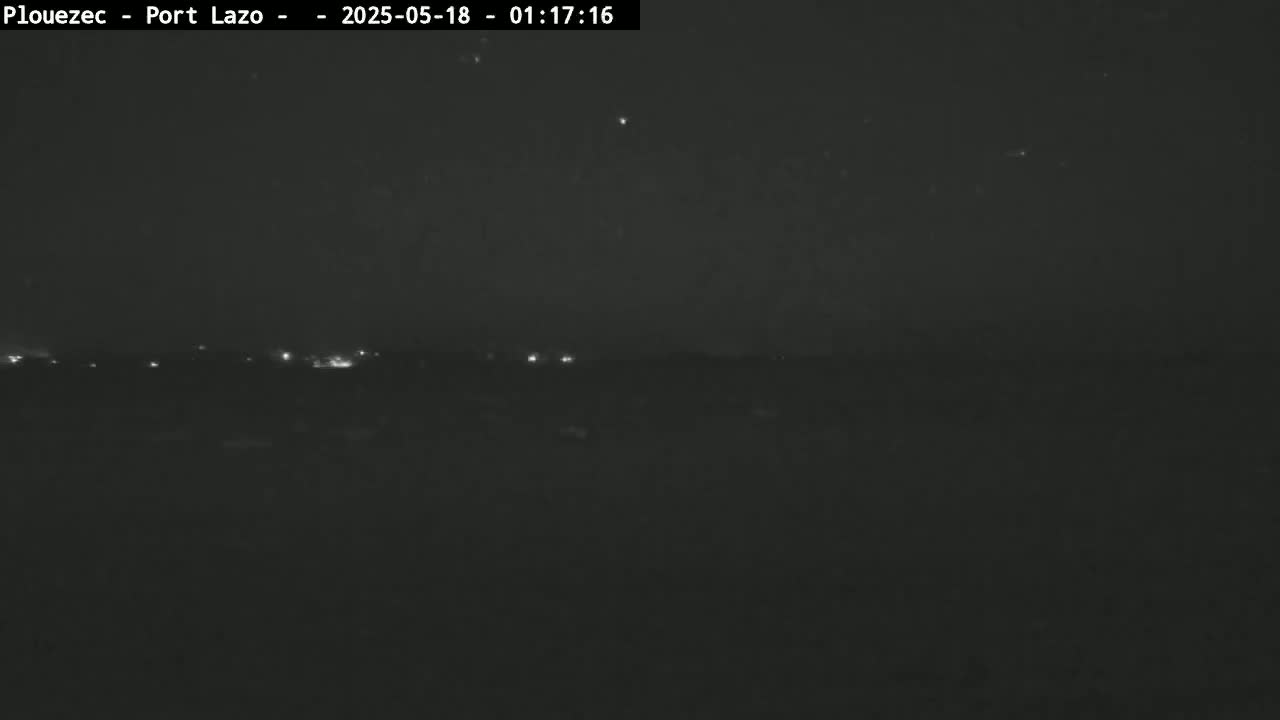 A nighttime, long shot of a dark landscape with a few faint lights visible in the distance under a clear, dark sky.