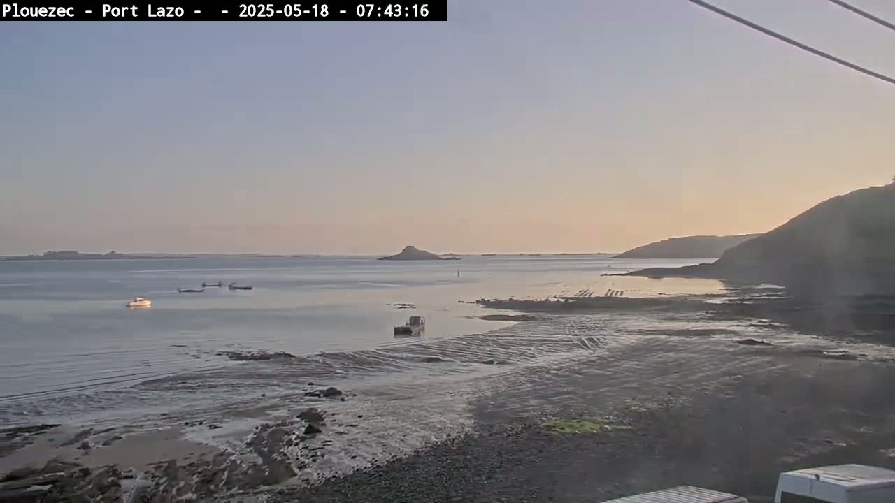 A calm, pale-sunset-lit bay shows several small boats on the water, a rocky shoreline, and a distant hill under a clear sky.