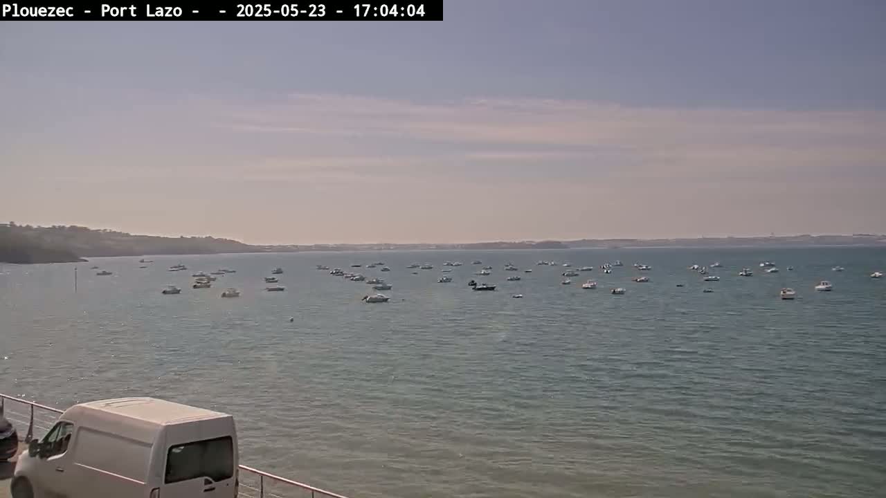 Numerous small boats are scattered across a calm body of water under a mostly clear sky, with a white van partially visible in the bottom left corner.