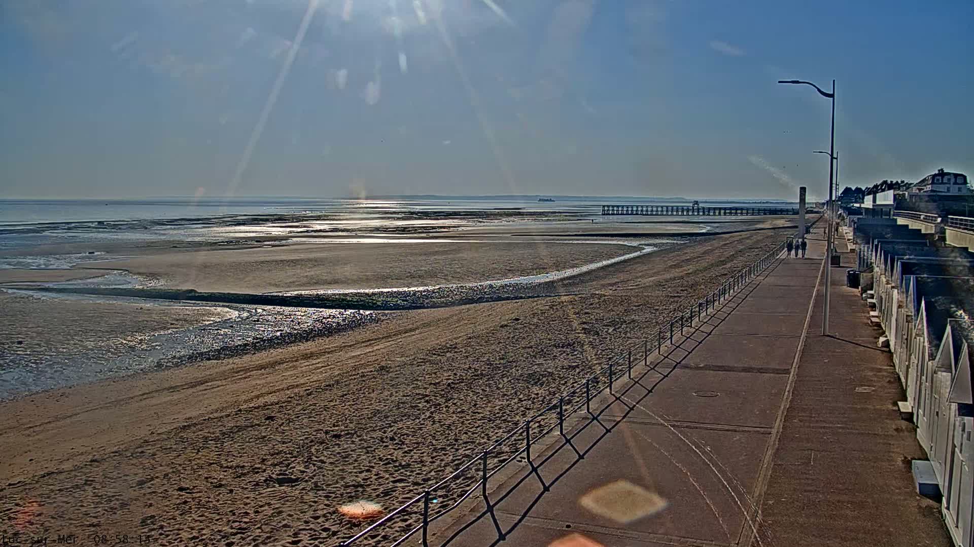 A sandy beach stretches to a calm sea under a bright, sunny sky, with a paved walkway and beach huts lining the shore.