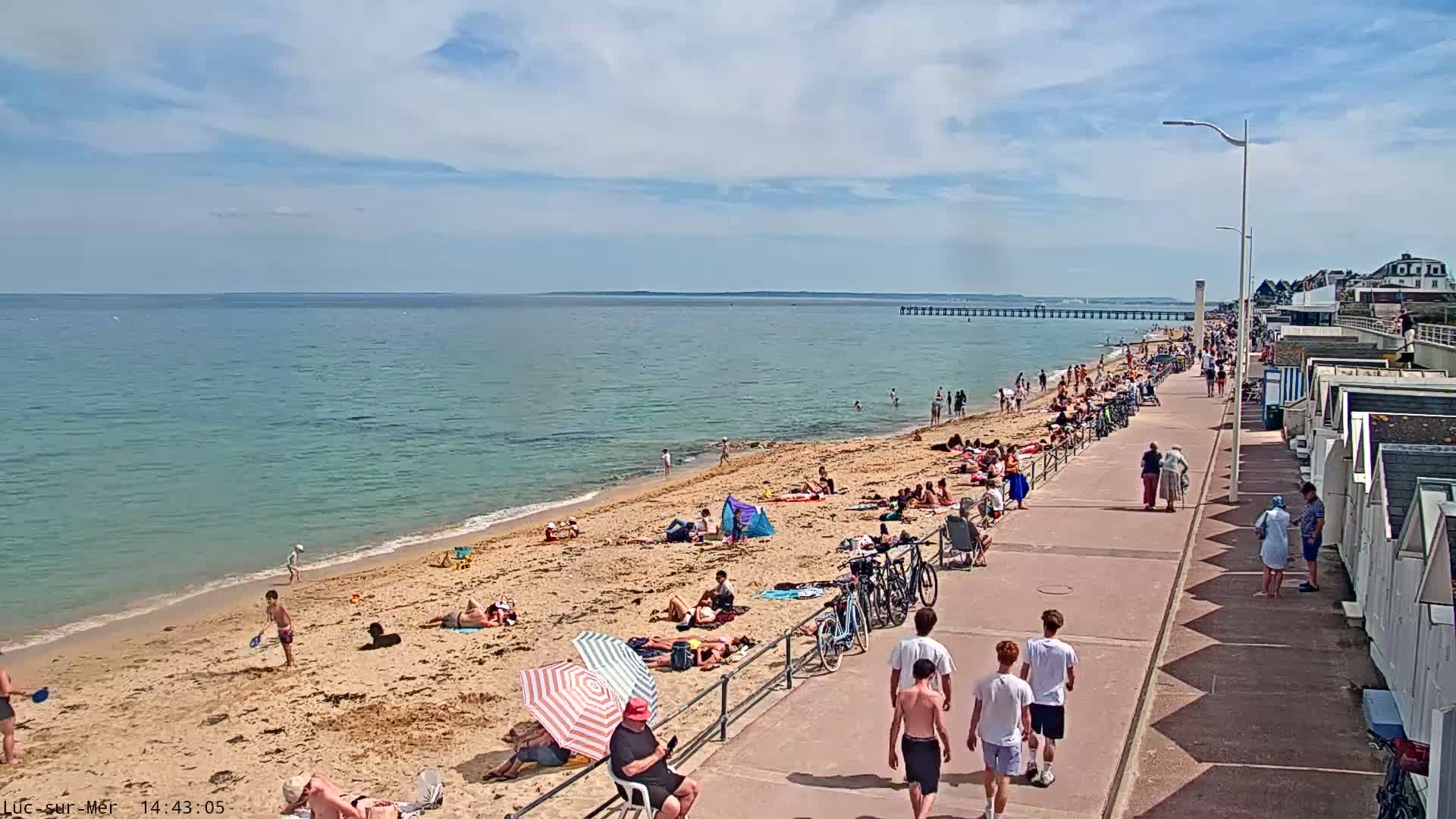 A sunny day at a crowded beach shows many people relaxing on the sand and walking along a boardwalk, with a pier visible in the distance.