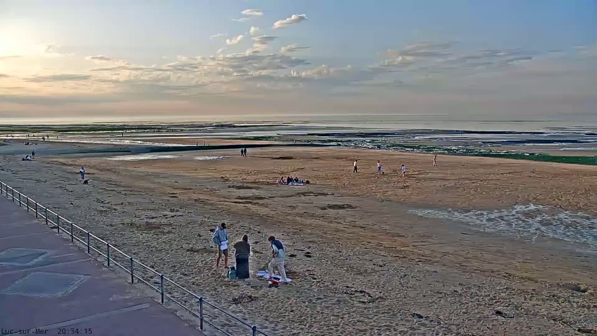 A sandy beach with scattered people and groups, under a partly cloudy sky, stretches to a calm ocean at low tide.