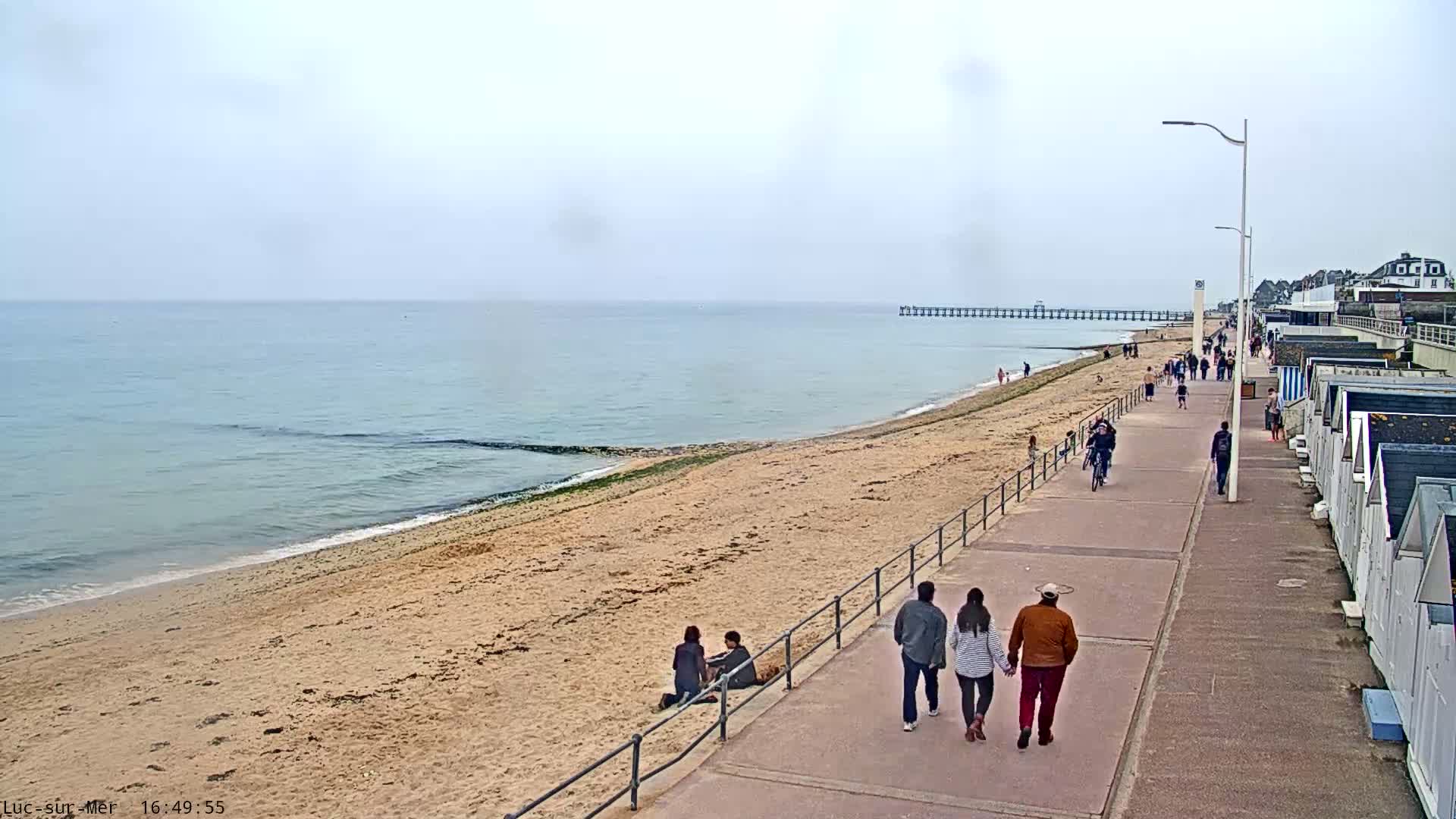 A sandy beach with people walking along a paved promenade next to a calm sea under an overcast sky.