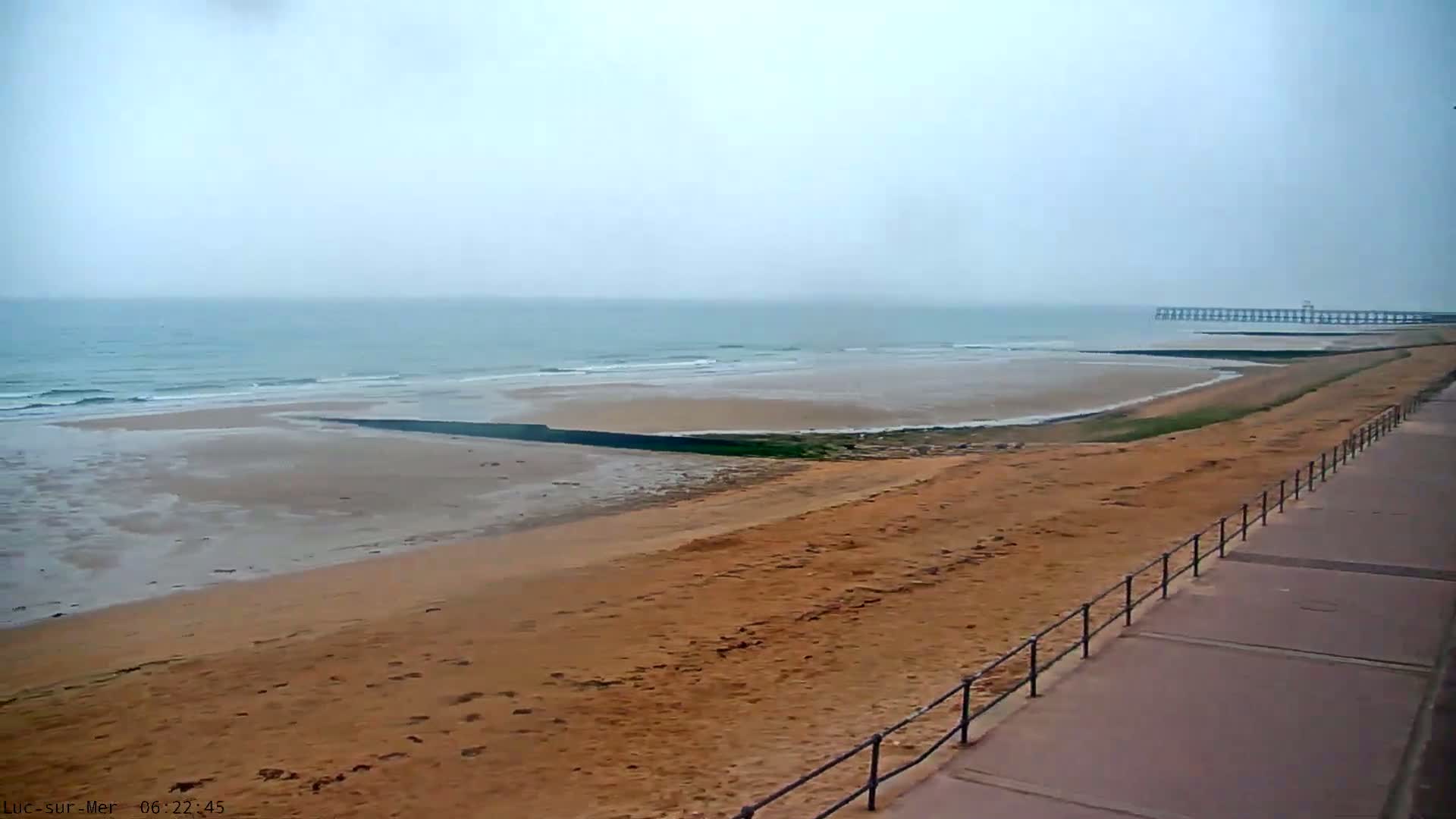 A long sandy beach with a boardwalk and a distant pier is visible under an overcast sky.