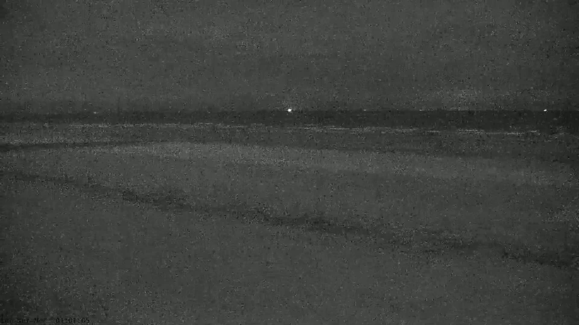 A nighttime, low-light image shows a dark beach and a dark ocean with faint lights visible on the horizon.