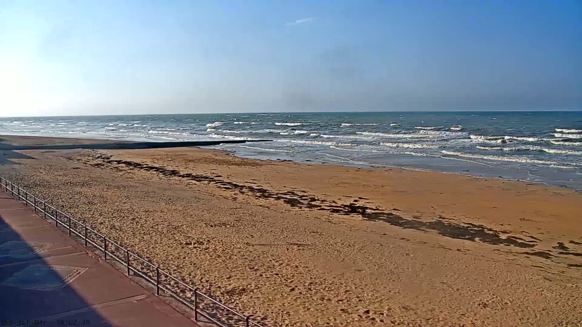 A sandy beach with a metal railing and a somewhat wavy ocean under a clear sky is visible.